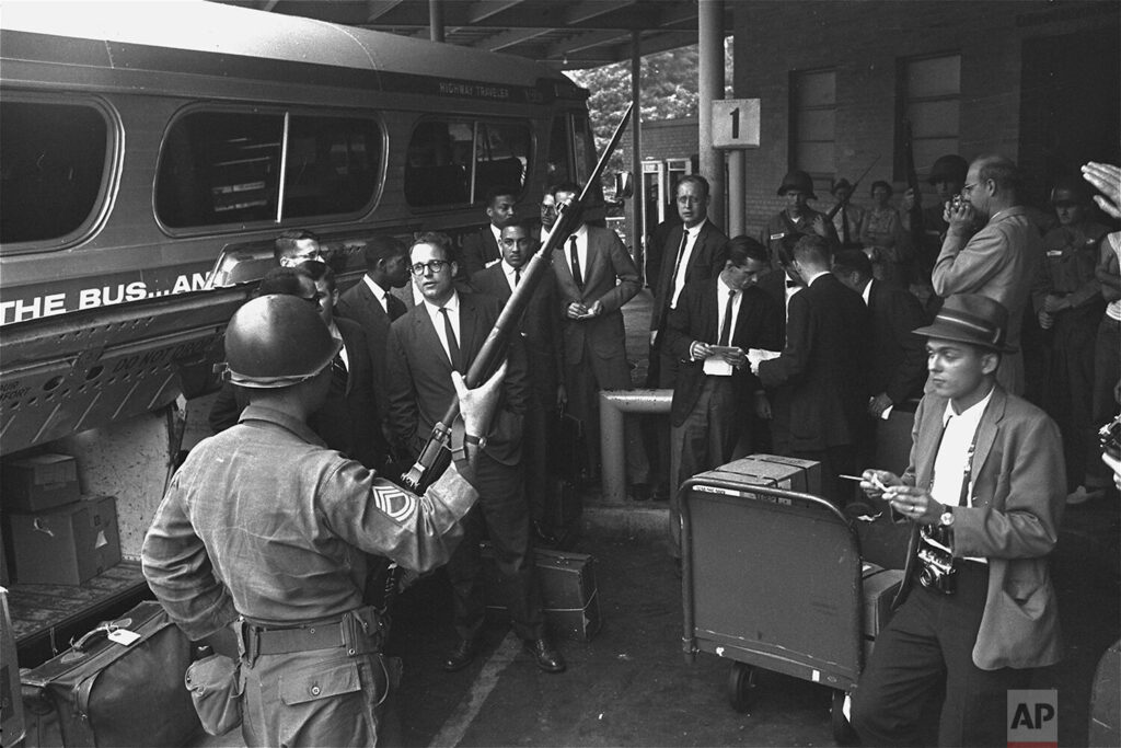 On the far left of the picture is a bus. Standing in front of the bus is a group of white professors and a racially diverse group of students, who were attempting to board. In the front of the picture if a member of the Alabama National Guard.