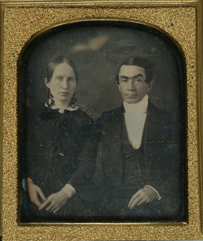 This is a daguerreotype of Eli Smith and his wife. Smith's wife is on the left, wearing an all-black outfit with a white collar and white sleeves that slip out. Smith is wearing a black suit in the 19th-century style with his left arm around his wife's back.